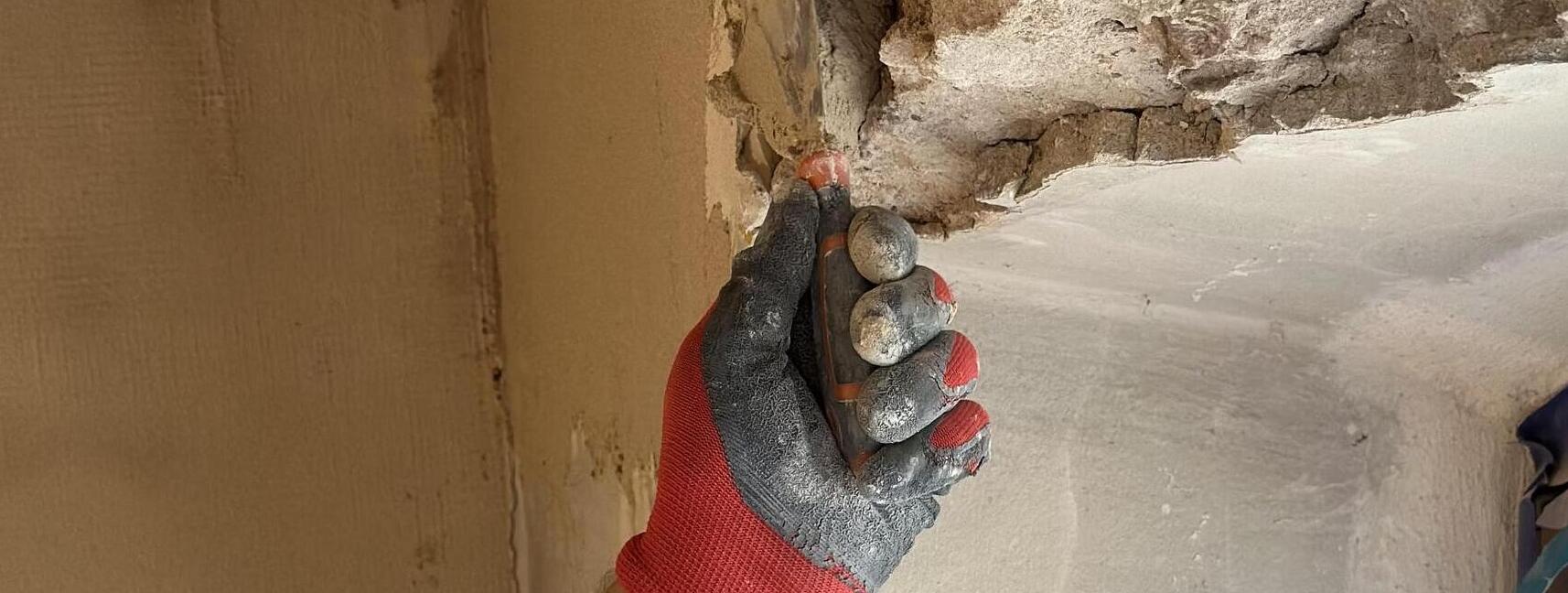 Hand im Arbeitshandschuh prüft beschädigten Putz an einer Wandecke